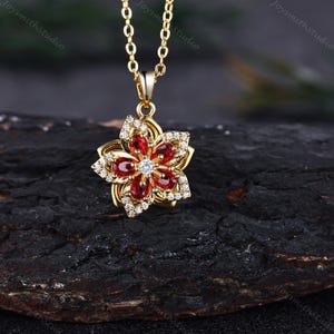 May include: A gold-toned necklace featuring a flower-shaped pendant. The pendant has five teardrop-shaped red stones arranged as petals, accented with small clear stones along the edges and a single clear stone at the center of the flower.