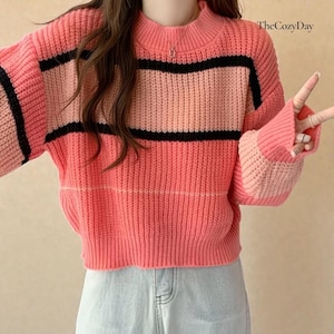 Op de afbeelding: Een roze, cropped trui met een hoge halslijn en lange mouwen. De trui heeft horizontale strepen in lichtroze, zwart en perzik. The Cozy Day staat rechtsboven afgedrukt.