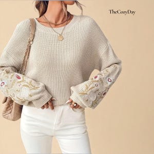 Könnte beinhalten: Ein cremefarbener Pullover mit floraler Stickerei an den Ärmeln. Der Pullover hat einen runden Ausschnitt und eine lockere Passform. Die Ärmel sind mit detaillierter Stickerei in Rot-, Gold- und Grüntönen versehen. The Cozy Day ist oben rechts aufgedruckt.