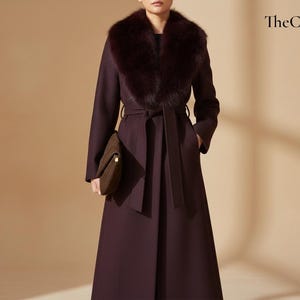 Puede incluir: Abrigo largo burdeos oscuro con cuello de piel sintética y cinturón a juego. El abrigo tiene un diseño clásico y se combina con un bolso marrón. The Cozy Day está escrito en la esquina superior derecha.