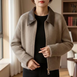 May include: A beige zip-up jacket with a brown leather collar. The jacket has ribbed cuffs and a ribbed hem. The model is wearing a black shirt underneath. The Cozy Day is written on the top right corner.