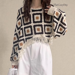 Op de afbeelding: Een gehaakte trui met een vierkant patroon in zwart, crème en bruin. De trui heeft lange mouwen, een ronde halslijn en een gefranjerde zoom. De tekst "TheCozyDay" staat in de rechterbovenhoek.