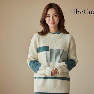 Könnte beinhalten: Cremefarbener Pullover mit tealfarbenen Akzenten. Der Pullover hat ein geometrisches Design auf der Vorderseite und den Ärmeln, mit vertikalen Streifen und Farbblöcken. Der Text "The Cozy Day" befindet sich oben rechts.