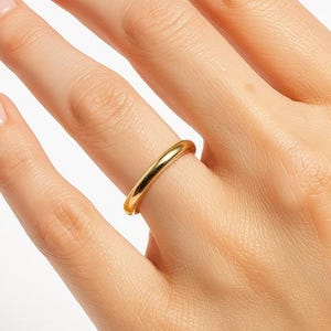 May include: A gold ring is worn on a finger. The ring is a simple band with a rounded top. The ring is a bright, shiny gold color. The skin tone of the hand is fair.