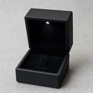 May include: A black jewelry box with a hinged lid, open to reveal a black velvet interior. A small LED light is positioned at the top. The box is designed to hold a ring.