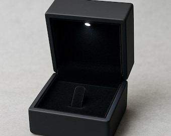 May include: A black jewelry box with a hinged lid, open to reveal a black velvet interior. A small LED light is positioned at the top. The box is designed to hold a ring.