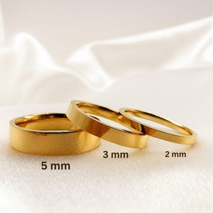 May include: Three gold-colored rings of varying widths: 2 mm, 3 mm, and 5 mm. The rings are displayed on a white, silky surface. The ring with a 5 mm width has a textured finish, while the others have a smooth, polished appearance. The rings are likely for a wedding or engagement.