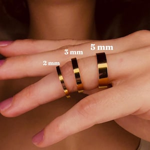 May include: Three gold-colored rings of varying widths: 2 mm, 3 mm, and 5 mm. The rings are displayed on a finger, showcasing their different sizes and a polished finish. The image highlights the rings' simple, elegant design.