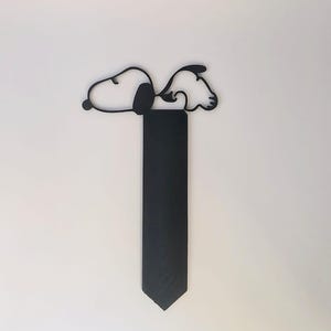 May include: A black metal Snoopy bookmark. The bookmark features a silhouette of Snoopy lying down on top of a rectangular black piece. The bookmark is designed to be inserted into a book to mark a page.