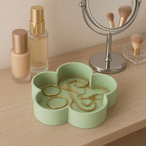 May include: A pale green, flower-shaped tray contains gold jewellery, including two hoop earrings and a delicate chain necklace. Two bottles of makeup and a small mirror are on the wooden surface. A makeup brush is visible.
