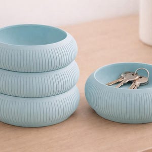 May include: Four light blue, ribbed, round catch-all trays. Three are stacked, and one holds keys. The trays are made of a solid material and sit on a light brown surface. The trays are a decorative storage solution.