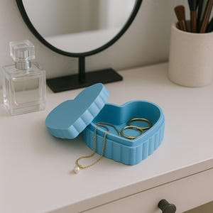 Pode incluir: Uma caixa de joias azul clara em forma de coração com um design canelado, aberta para revelar anéis dourados e um colar com um pingente de pérola. Um frasco de perfume e um espelho estão no fundo.