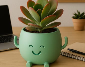 Puede incluir: Una maceta de cerámica verde con una cara sonriente, brazos y piernas, que contiene una planta suculenta con hojas verdes y rojas. La maceta está sobre un escritorio de madera, con un portátil, un reloj y un bloc de notas en el fondo.