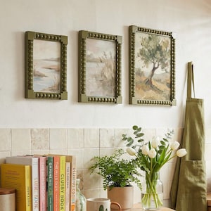 May include: A kitchen interior featuring framed landscape paintings, a cactus-themed mug, and a coffee machine. The room includes a wooden counter with books and plants, a stainless steel toaster, and a green apron. The style is rustic and welcoming.