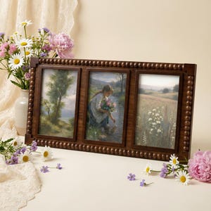 Peut inclure: Un cadre photo triple en bois brun foncé avec une bordure perlée. Chaque cadre contient une peinture de paysage différente. Les peintures représentent un arbre, une femme près d'un ruisseau et un champ de fleurs. Le cadre est présenté avec des fleurs.