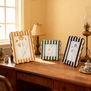 Puede incluir: Tres marcos de fotos decorativos con bordes festoneados y patrones de rayas. Los marcos muestran obras de arte florales. Uno tiene rayas amarillas y blancas, otro verdes y blancas, y el tercero negras y blancas. Están colocados sobre un escritorio de madera.