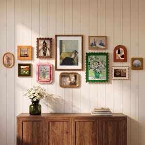 May include: A gallery wall arrangement featuring twelve assorted picture frames in various shapes, sizes, and colors, including scalloped, rectangular, and oval designs, displayed on a white paneled wall above a wooden cabinet with a vase of daisies.