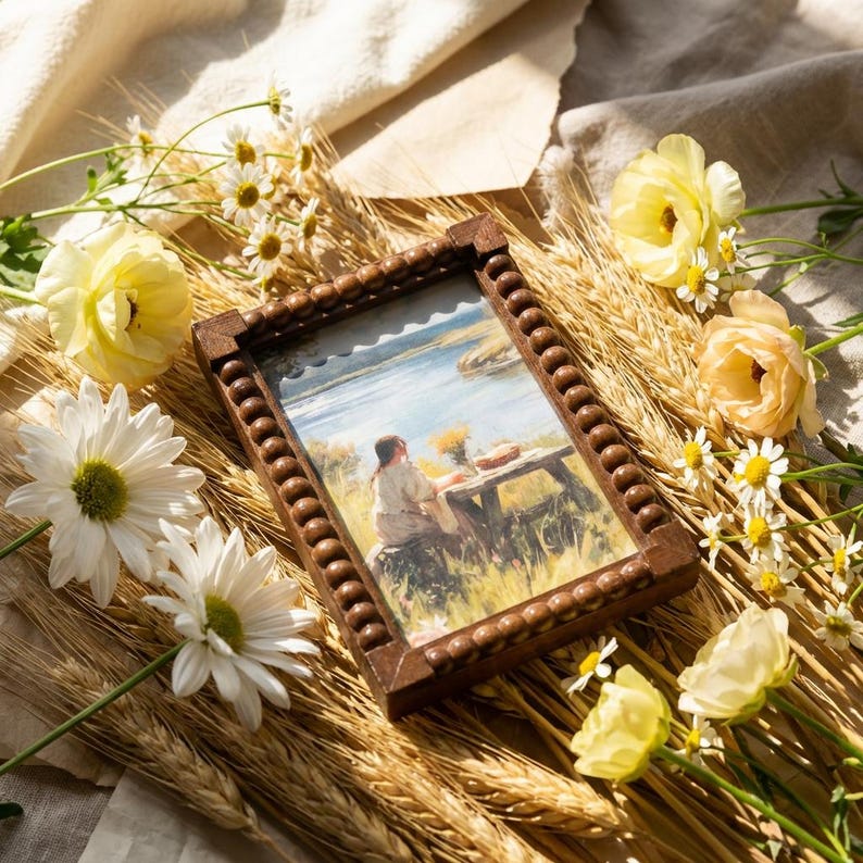 May include: A rectangular wooden picture frame with a beaded border containing an illustration of a person sitting at a table in a field near water. The frame is surrounded by yellow ranunculus flowers, white daisies, and dried wheat stalks.