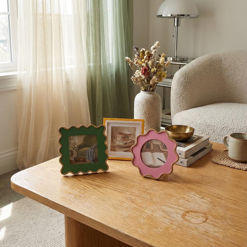 May include: Three decorative picture frames with scalloped edges in green, yellow, and pink sit on a wooden table. A vase of dried flowers, a brass bowl, and a mug are also on the table. Sheer curtains are in the background.