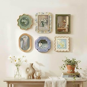May include: A gallery wall arrangement featuring six decorative frames with various art prints, including floral motifs, a swan, and a glass of lemonade. Below, a wooden table holds a glass vase with white flowers, a ceramic vase, and a candle.