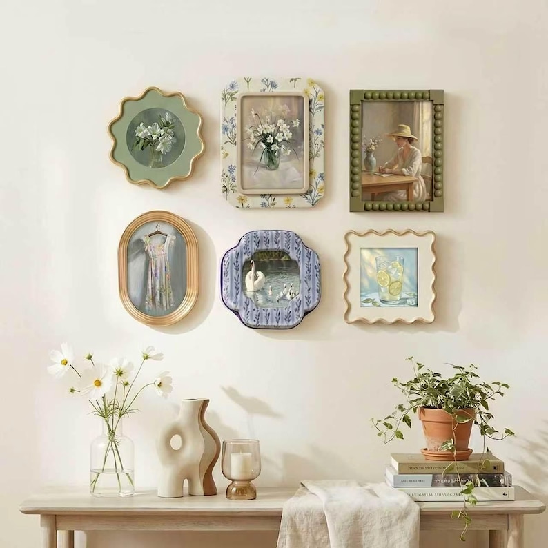 May include: A gallery wall arrangement featuring six decorative frames with various art prints, including floral motifs, a swan, and a glass of lemonade. Below, a wooden table holds a glass vase with white flowers, a ceramic vase, and a candle.
