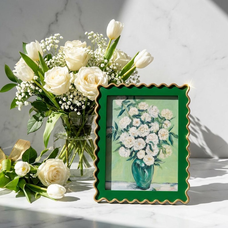 May include: A rectangular picture frame with a dark green scalloped border and a gold inner edge, containing a painting of white flowers in a green vase. The frame is positioned next to a glass vase filled with white roses and baby's breath.