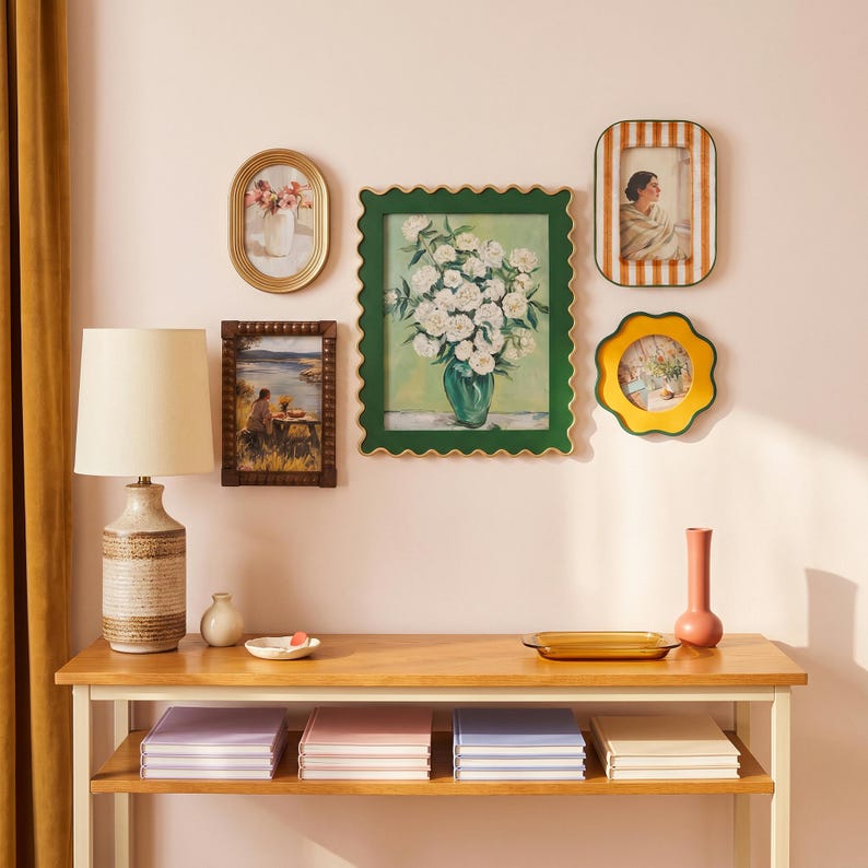 May include: A gallery wall arrangement featuring five assorted frames, including a large scalloped green frame with a floral painting, a gold oval frame, a dark wood frame, a striped frame, and a yellow flower-shaped frame above a wooden console table.