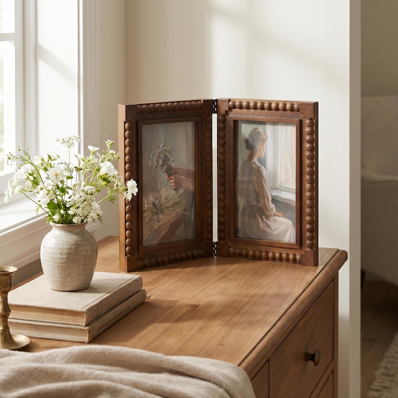 May include: A hinged double picture frame made of dark brown wood with a beaded border design. The frame holds two rectangular prints, one depicting a person holding flowers and the other showing a person sitting by a window, both in soft tones.