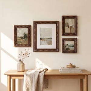 Puede incluir: Cuatro pinturas de paisajes enmarcadas con marcos de cuentas marr&oacute;n oscuro se exhiben en una pared de color crema. Las pinturas representan escenas con colores apagados, incluyendo verdes, marrones y blancos. Una mesa de madera con un jarr&oacute;n, libros y un cuenco de madera est&aacute; en primer plano.