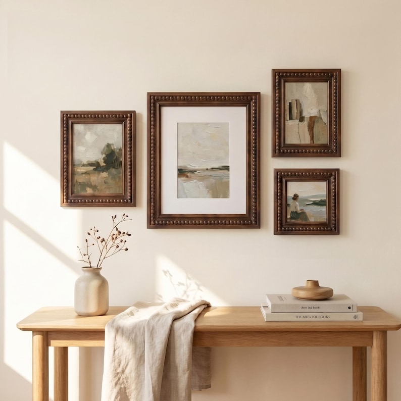 Puede incluir: Cuatro pinturas de paisajes enmarcadas con marcos de cuentas marr&oacute;n oscuro se exhiben en una pared de color crema. Las pinturas representan escenas con colores apagados, incluyendo verdes, marrones y blancos. Una mesa de madera con un jarr&oacute;n, libros y un cuenco de madera est&aacute; en primer plano.