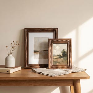 Puede incluir: Dos pinturas de paisajes enmarcadas con marcos marrones con cuentas descansan sobre una mesa de madera. Un peque&ntilde;o jarr&oacute;n blanco con flores secas y un libro cerrado tambi&eacute;n est&aacute;n sobre la mesa. Un pa&ntilde;o de lino doblado se coloca frente a las pinturas.