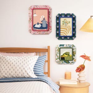 Könnte beinhalten: Drei dekorative gerahmte Kunstwerke mit floralen und geometrischen Mustern hängen an einer weißen Wand. Die Rahmen zeigen farbenfrohe Kunstwerke von Stillleben. Ein Bett mit Kissen und ein Beistelltisch mit einer Kerze und einer Vase befinden sich im Vordergrund.