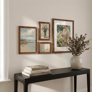May include: A collection of framed art pieces with wooden beaded frames, including landscape and portrait paintings. A vase with dried branches sits on a black console table, with a stack of books below the art.
