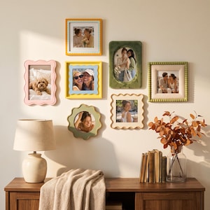 May include: A gallery wall arrangement featuring seven assorted picture frames in various shapes and colors, including scalloped, beaded, and smooth edges in yellow, pink, green, and cream, displaying photographs of people and a dog.