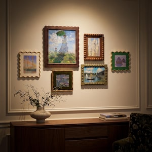 May include: A gallery wall arrangement featuring six framed art prints in various styles, including scalloped and beaded frames, displayed above a wooden sideboard with a ceramic vase containing branches and a stack of books.