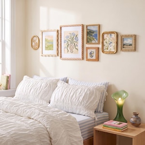 May include: A gallery wall featuring seven assorted framed art prints with gold and textured frames above a bed with white ruched bedding, light blue striped sheets, a green glass lamp, and a small ceramic vase on a wooden nightstand.