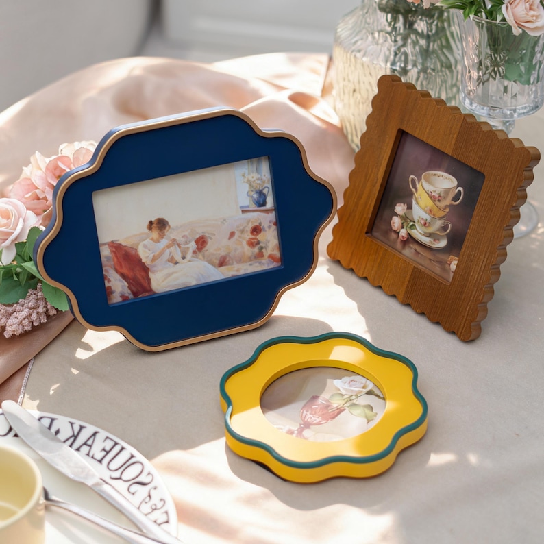 May include: Three decorative picture frames in different styles. A navy blue frame with a scalloped edge, a wooden frame with a square image, and a yellow frame with a scalloped edge. The frames are on a table with a soft pink tablecloth.