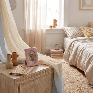May include: A bedroom scene featuring a light pink picture frame with a floral arrangement, two small vases, and a patterned cloth on a white wooden chest. Soft, flowing curtains in cream and blue hues frame the window and bed with neutral-toned bedding.