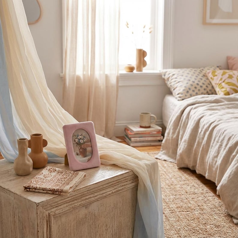 May include: A bedroom scene featuring a light pink picture frame with a floral arrangement, two small vases, and a patterned cloth on a white wooden chest. Soft, flowing curtains in cream and blue hues frame the window and bed with neutral-toned bedding.