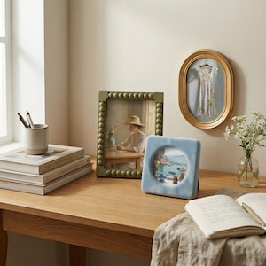 May include: A collection of home decor items including a rectangular olive green beaded picture frame, an oval gold picture frame, and a square light blue velvet picture frame. Also featured are a ceramic pen holder and stacked books.