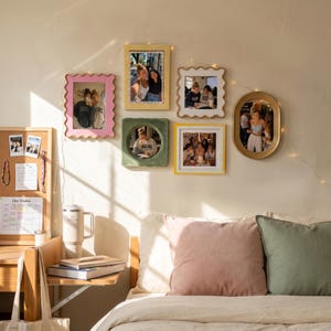 May include: A collection of six wall-mounted picture frames in various shapes and colors, including pink, yellow, green, white, and gold, arranged around a string of fairy lights above a bed with pink and green pillows and a cork board.