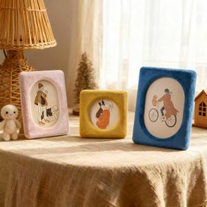 Può includere: Tre cornici per foto in peluche rosa, giallo e blu. Ogni cornice ha un'apertura ovale con un disegno all'interno. La cornice rosa mostra una persona che porta a spasso un cane. La cornice gialla mostra una persona che tiene un gatto. La cornice blu mostra una persona in bicicletta.