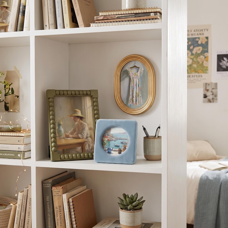 May include: A white bookshelf containing a green beaded picture frame with a portrait, an oval gold frame displaying a miniature dress, a square light blue velvet frame with a coastal print, and a two-toned ceramic cup holding two pens.