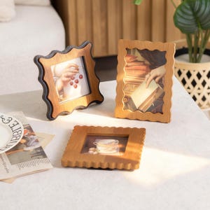 May include: Three wooden picture frames with a scalloped design. Two frames are light brown, one with a dark brown edge. The frames display images of hands, books, and a teacup. The frames are on a white surface.