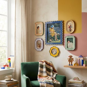 May include: A collection of framed art pieces in various shapes and sizes, mounted on a wall. The frames are in shades of blue, yellow, and white, with images of people and flowers. A green armchair with a striped blanket is in the foreground.