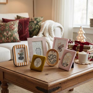 Pode incluir: Uma coleção de molduras decorativas em várias formas e cores, incluindo rosa, amarelo e listras. As molduras exibem estampas botânicas e artísticas, dispostas sobre uma mesa de centro de madeira. Um ambiente aconchegante de sala de estar com almofadas e uma árvore de Natal.