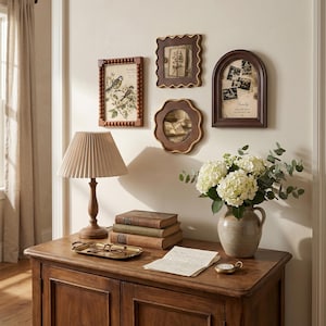May include: A wooden console table holds a pleated lamp, a stack of three books, a gold tray with glasses, a ceramic vase with white hydrangeas, and a pocket watch. Four wall frames display botanical art, family photos, and text.