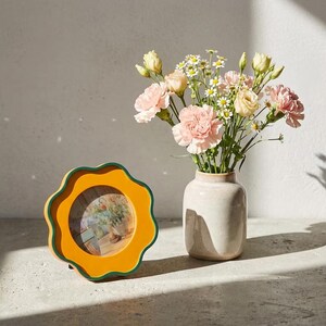 May include: A circular picture frame with a wavy, scalloped edge in mustard yellow and dark green, positioned next to a light beige ceramic vase filled with a bouquet of pink carnations, pale yellow lisianthus, and small white daisies.