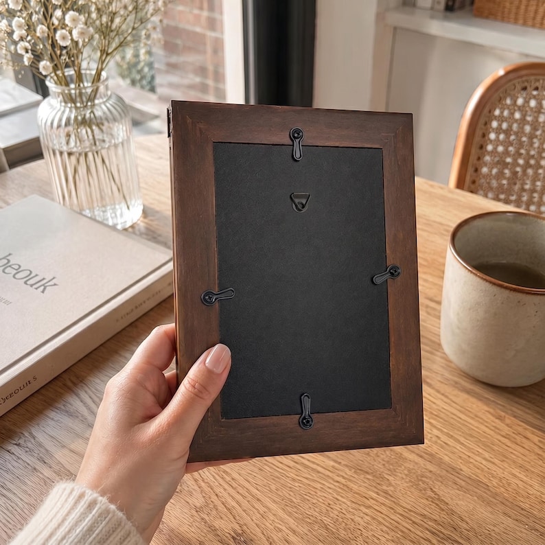 May include: A rectangular dark brown wooden picture frame shown from the back. The frame features a black backing board secured by four black metal swivel clips and includes a central black metal triangular hanging hook for wall mounting.