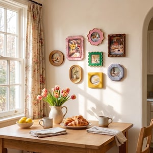 May include: A collection of seven decorative wall frames in various shapes and colors, including pink, green, yellow, and wood tones, arranged on a wall above a wooden dining table set with a ceramic pitcher of tulips, croissants, and mugs.